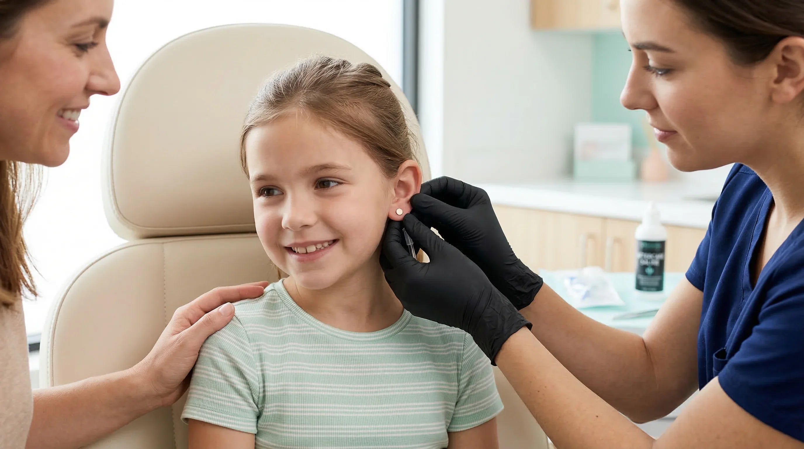Glückliches Mädchen beim Ohrlochstechen durch Fachkraft mit schwarzen Handschuhen, Mutter beruhigt sie.